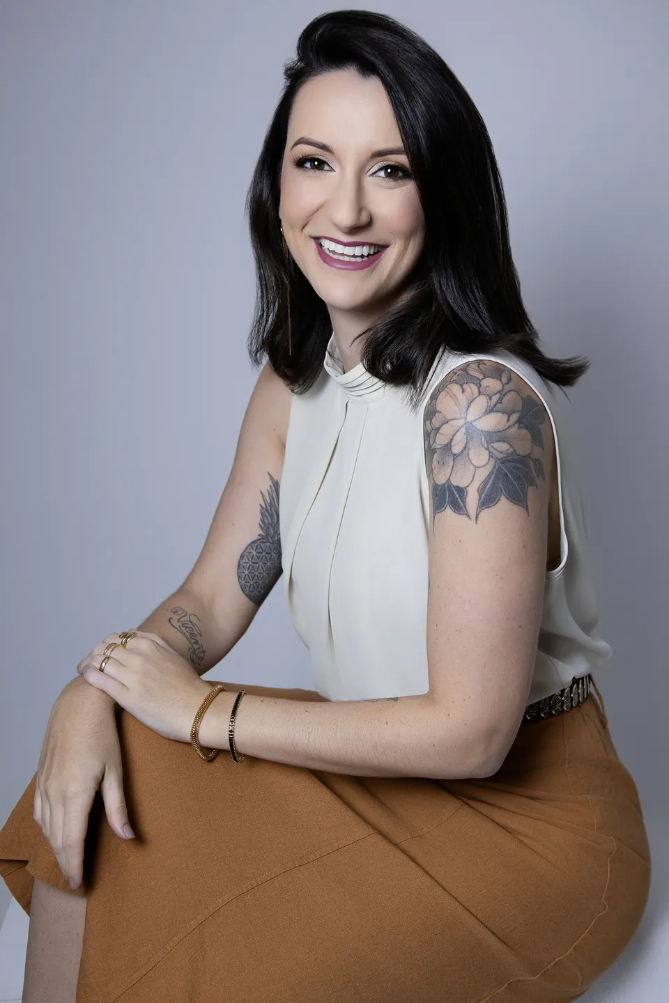 Julia Poltronieri sentada, sorrindo, com blusa clara e saia caramelo